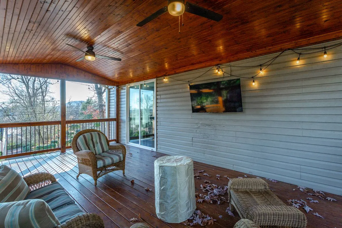A cozy outdoor deck featuring comfortable seating grouped around a flat-screen TV mounted on the wall, enhanced by string lights and a warm wooden ceiling. The space invites relaxation and entertainment, providing ample room for gatherings.