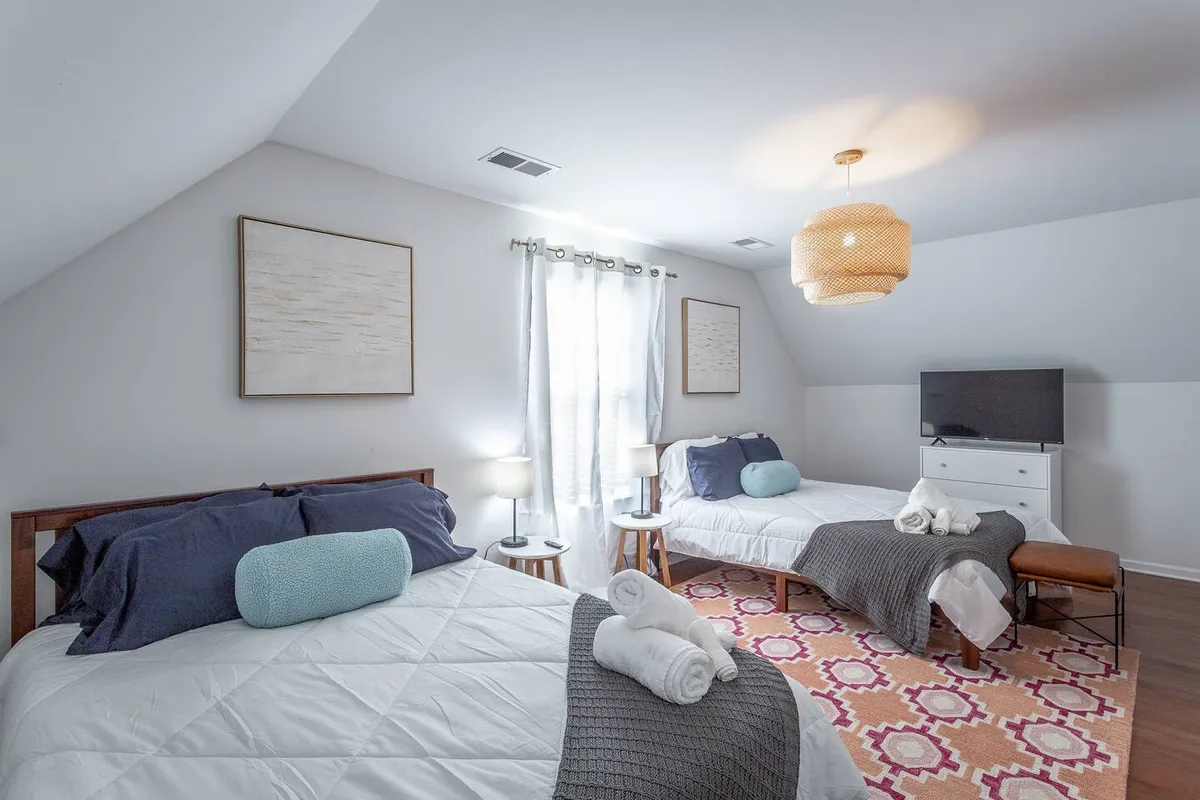 A bright bedroom with two beds topped with decorative pillows, a colorful area rug, a large window letting in natural light, and a modern light fixture overhead. The space feels fresh, comfortable, and well-suited for groups or families.