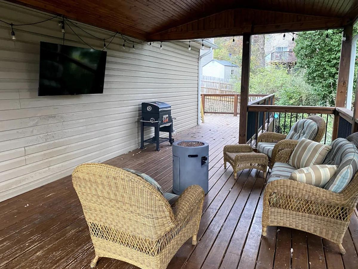 This exterior shot features a cozy deck furnished with wicker chairs, a fire pit, a grill, and a flat-screen TV mounted outside. The setup invites outdoor gatherings and movie nights under the stars, surrounded by a relaxed, welcoming atmosphere.