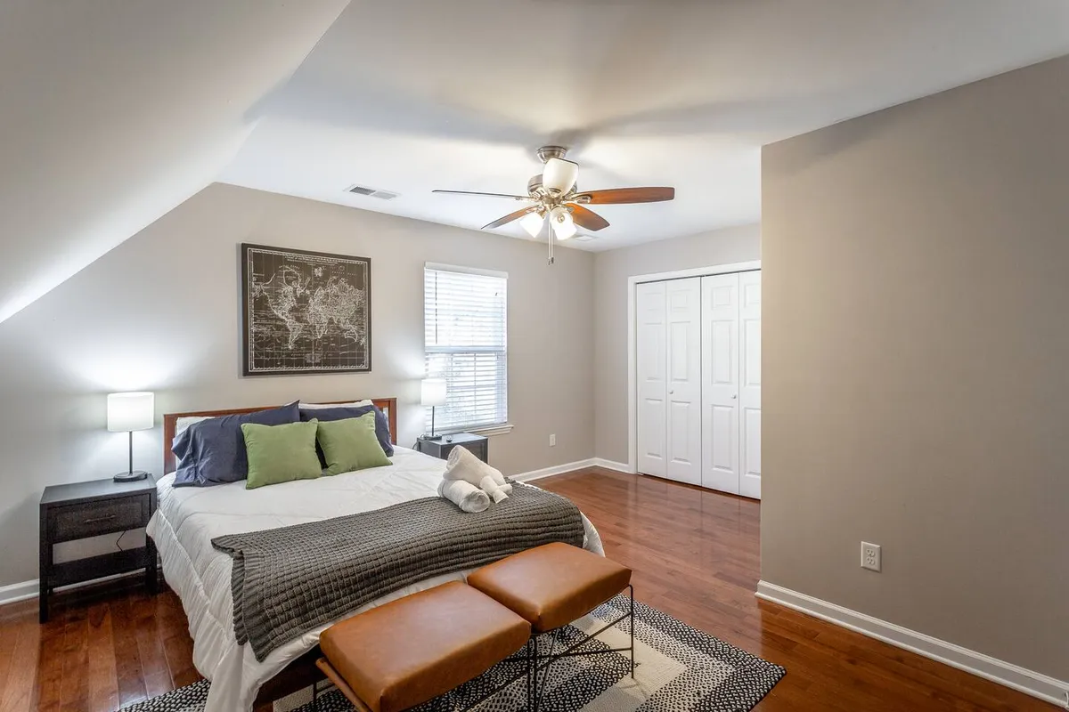 A serene bedroom featuring a bed with decorative pillows, a framed map on the wall, a nightstand with a lamp, and two ottomans at the foot of the bed. Soft lighting gives the space a calm, welcoming feel, ideal for relaxation and private reflection.