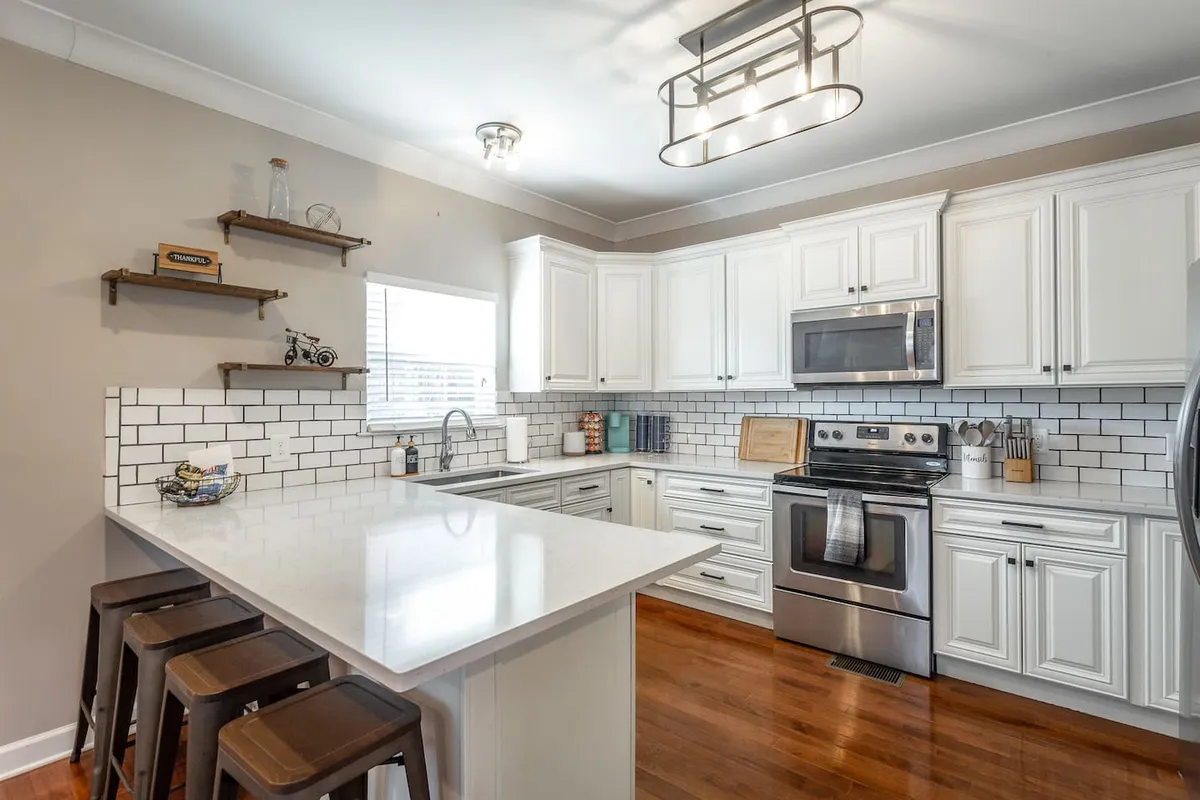 A modern kitchen featuring a spacious island, white cabinetry, a stylish light fixture above, and stainless steel appliances. The room is open, with clear sightlines to adjoining living and dining areas, inviting both the cook and guests to mingle easily.