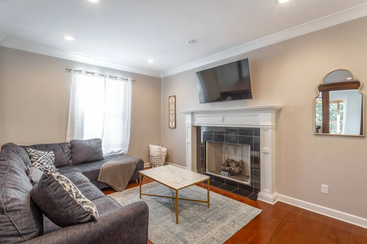 A cozy living room with a sofa adorned with decorative pillows, a coffee table, a fireplace, a mounted television, and a window letting in natural light. The arrangement is welcoming and encourages relaxation or socialization.
