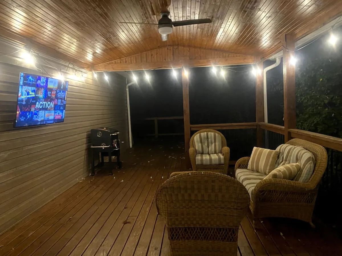 An inviting exterior seating area on a deck, featuring wicker chairs, a grill, a large television screen, and string lights overhead, ideal for group socializing, barbecues, and outdoor movie nights under the stars.