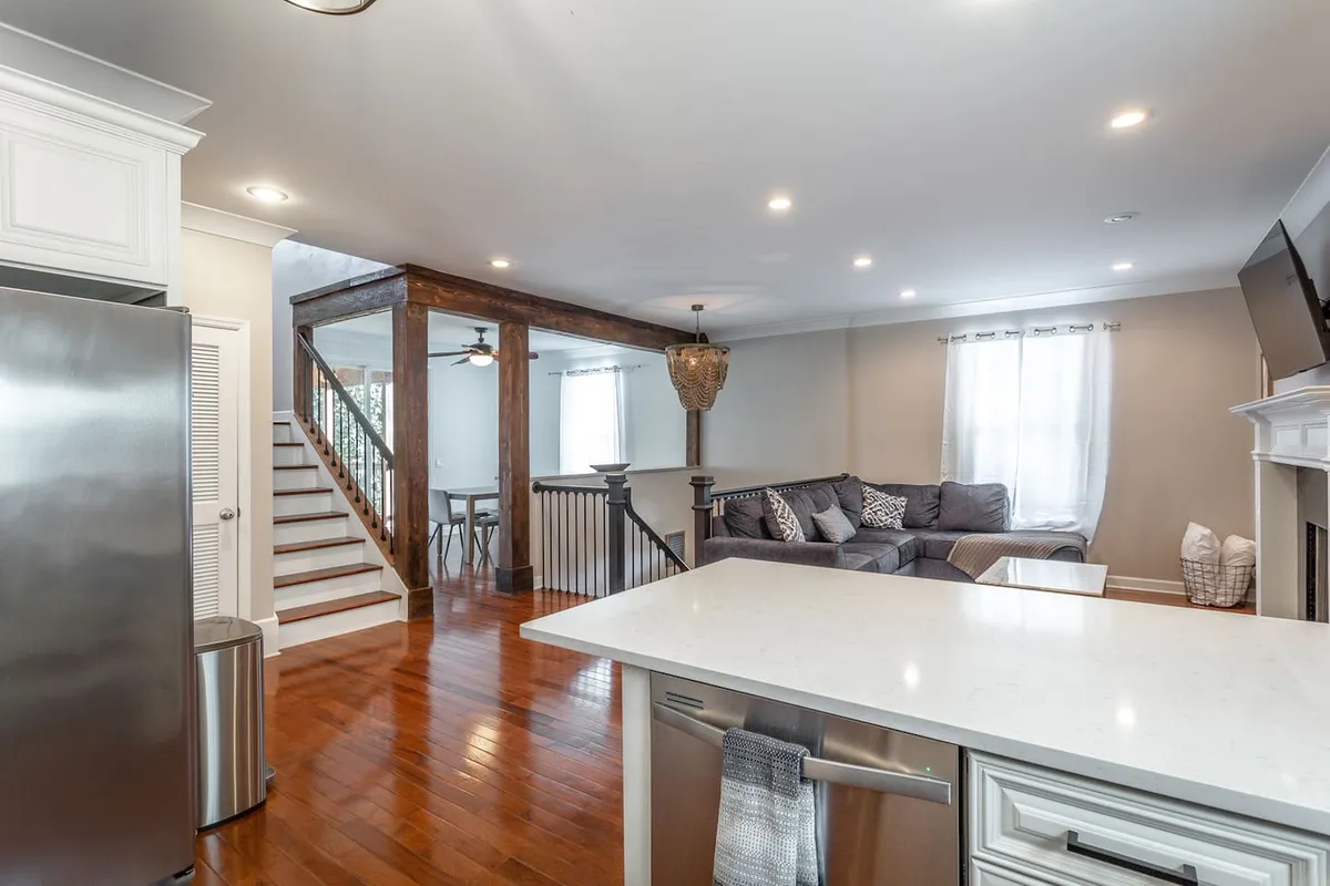 A spacious interior featuring a kitchen island, a comfortable sofa, a prominent staircase, and a dining area arranged for a group. The room is well-lit, with stylish decor and furnishings that encourage gathering and conversation.